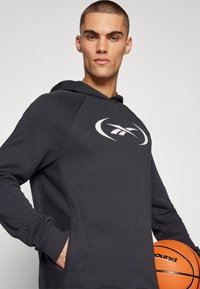 Sweat à capuche gris foncé avec une finition texturée et un logo blanc. Le modèle tient un ballon de basket orange, mettant en avant l'ajustement décontracté du vêtement.