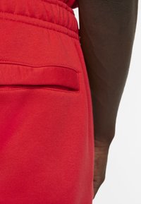 Red cotton joggers with an elastic waistband, featuring a back pocket and smooth texture. Close-up of fabric and pocket detail.