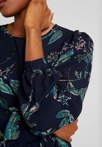 Marinblå blommig klänning med puffärmar, med gröna löv och rosa blommor. Tyget har en mjuk textur med en subtil lyster.