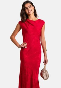 Abito rosso con motivo floreale, collo a barca e maniche a cappuccio. La modella tiene una piccola borsa beige, accessoriate con bracciali dorati.