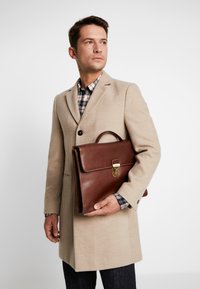Maletín de cuero marrón con un mango superior y cierre de metal, sostenido por una persona con un abrigo beige sobre una camisa de cuadros. Diseño sencillo.
