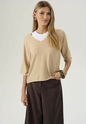 Giovane donna con lungo capelli biondi, che indossa un maglione beige a scollatura a V sopra una maglietta bianca, pantaloni larghi neri, orecchini a cerchio e un bracciale nero, con una mano in tasca.