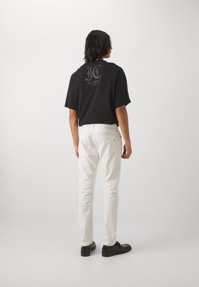 T-shirt nera con un logo sulla schiena, abbinata a jeans slim-fit bianchi. Le scarpe sono nere e hanno una finitura lucida.