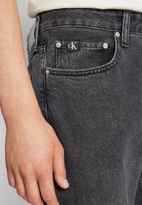 Jean en denim gris foncé avec une texture subtile, design à cinq poches et un logo cK noir sur la poche avant, agrémenté de détails en métal argenté.