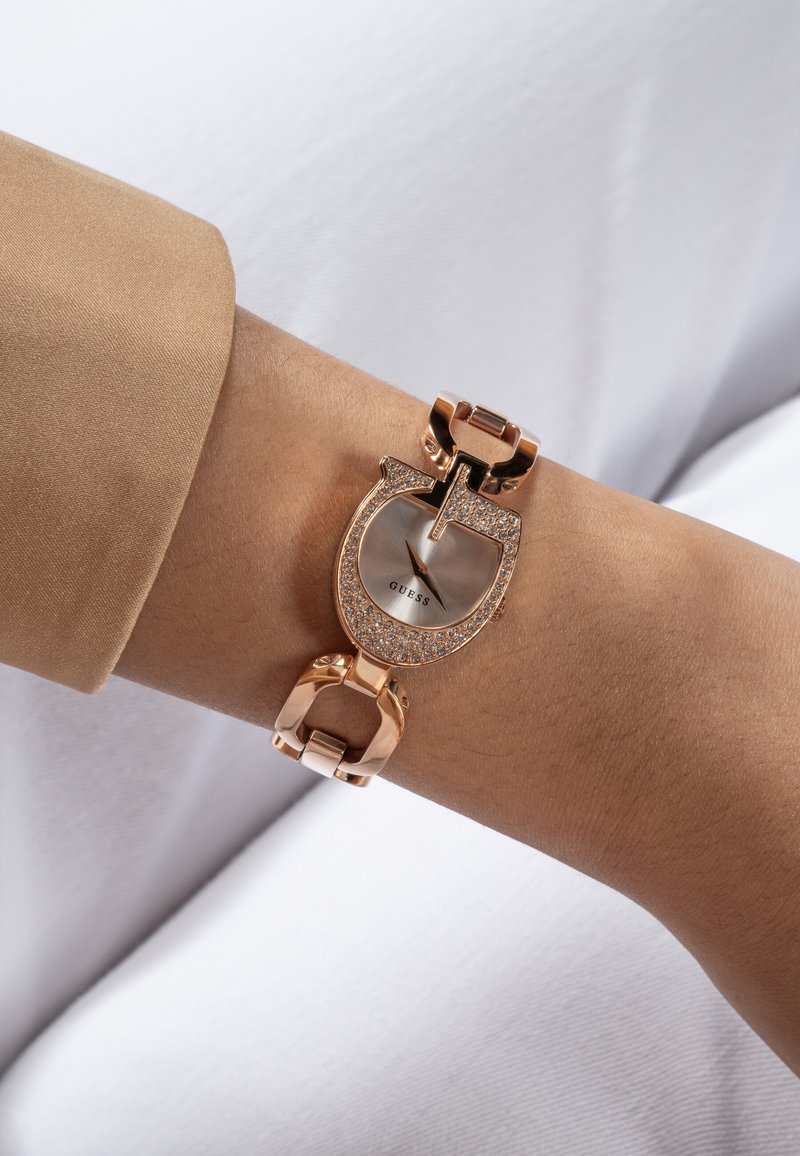 Roza zlata ura Guess z bleščečo ohišjem v obliki konjskega zanke in verižnimi povezavami na zapestju, oblečenem v bež rokav.