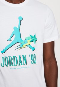 Camiseta de algodón blanca con un gráfico turquesa de un jugador de baloncesto, el texto "JORDAN '92" y una estrella con un diseño dinámico.
