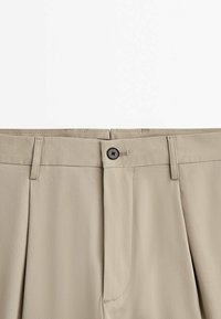 Pantalon beige vu de face avec taille boutonnée, passants de ceinture et plis cousus, présenté sur fond blanc uni.