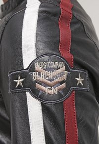 Sort læderjakke med hvide og røde striber. Har et rundt broderet mærke med stjerner og tekst, som tilføjer tekstur og detaljer.