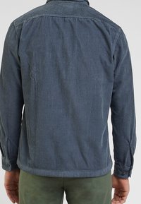 Giacca in corduroy blu scuro con texture a strisce verticali, maniche lunghe e polsini con bottoni. La vista posteriore mostra il colletto e l'orlo arrotondato.