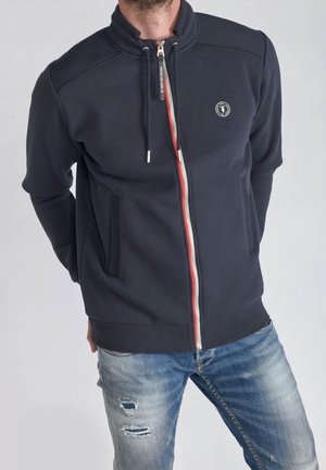 Homme portant une veste zippée bleu marine avec cordons et un logo rond sur la poitrine, associée à un jean bleu clair déchiré.