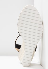 Semelle d'une sandale compensée blanche avec des rainures horizontales, une bride de cheville noire et un talon plateforme tissé texturé sur fond blanc.