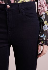 Jeans de mezclilla de tiro alto en color navy con una textura suave, con un bolsillo frontal y cierre de botón metálico. Mano parcialmente visible con una manga floral.