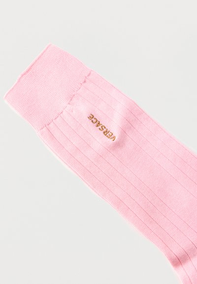 Ροζ ανοιχτό ριμπ κάλτσα με διπλωμένη μανσέτα, διακοσμημένη με χρυσή κεντητική "VERSACE" κοντά στην κορυφή, σε απλό λευκό φόντο.