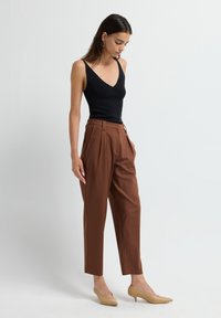 Vrouw in een zwarte mouwloze V-hals top, hoog-{waist} bruine plooienbroek, beige lage hakken schoenen, staande tegen een effen lichte achtergrond.