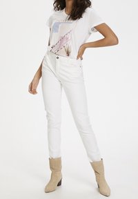 Mujer que lleva jeans blancos de tiro alto, botas de tacón de ante beige y una camiseta gráfica con el texto "POSITIVO" y un diseño de escalera en tonos pastel.