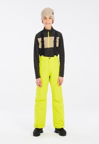 Gelbe wasserdichte Skihose, schwarzes Langarmshirt mit beigem Muster, graue Mütze und schwarze Schuhe. Verstellbare Träger inklusive.