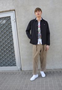 Černá bunda se cvoky, bílá kvetinová košile pod ní, béžové kordové kalhoty a bílé slip-on boty. Stojí opřený o betonovou zeď.