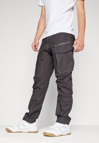 Nákladní kalhoty z tmavě šedé látky s několika kapsami na zip, se zúženým střihem a kontrastním prošíváním. Nošeny s bílými teniskami.