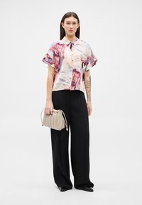 Žena s dlouhými tmavými vlasy, oblečená v tričku s krátkým rukávem a abstraktním potiskem, černé kalhoty širokých nohavic, drží béžovou kabelku s nařasením.