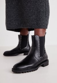 Bottines en cuir noir avec des panneaux latéraux en élasthanne, des clous décoratifs le long des bords et une semelle en caoutchouc texturée pour une bonne adhérence.
