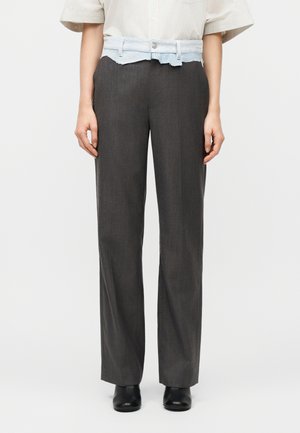 Kobieta w ciemnoszarych spodniach o prostym kroju, pod spodem warstwowo nosząca krótką niebieską dżinsową spódniczkę z surowym wykończeniem, w połączeniu z czarnymi butami.