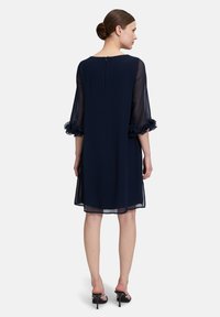 Robe bleu marine avec des volants transparents à manches 3/4 et une ouverture en clé à l'arrière. Tissu lisse, longueur genou, coupe décontractée.