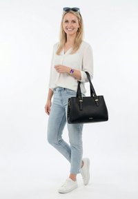 Smilende kvinde med langt blondt hår iført en hvid bluse, lyse jeans, hvide sneakers, solbriller på hovedet, lilla ur og holder en sort håndtaske.