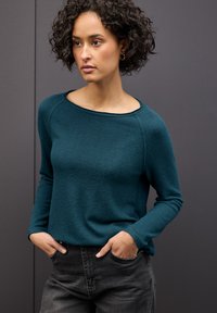 Türkisfarbener, strukturierter Pullover mit weitem U-Ausschnitt und langen Ärmeln, kombiniert mit dunkelgrauen Jeans. Das Model steht mit den Händen in den Taschen.