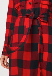 Robe à carreaux rouge et noire avec nœud à la taille et poches poitrine boutonnées, portée par une personne avec la main gauche visible.