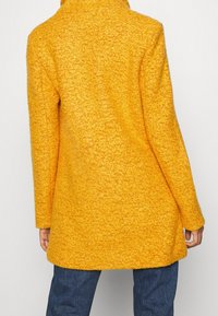 Abrigo texturizado amarillo con cuello alto, mangas largas y corte recto, combinado con vaqueros oscuros de mezclilla. Vista trasera muestra hardware mínimo.