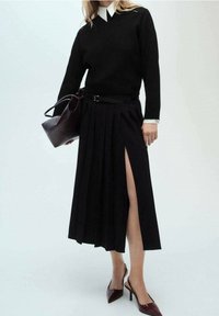 Femme portant un haut noir à manches longues avec col blanc, une jupe plissée noire fendue à la cuisse, tenant un sac à main noir, et des chaussures à talons bordeaux.