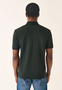 Ciemnozielona koszulka polo z krótkimi rękawami, kołnierzykiem i gładką teksturą, zestawiona z niebieskimi dżinsami. Widok od tyłu ukazuje linię dołu i dopasowanie.