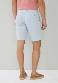 Man med ljusblå kavajshorts, rosa t-shirt och beige slip-on skor, stående mot en enhetlig ljus bakgrund.