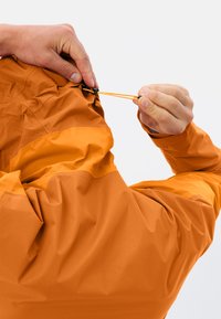 Giacca outdoor arancione con tessuto testurizzato, dotata di un cordino regolabile al collo tramite un fermacorda nero, che mette in evidenza elementi di design pratici.