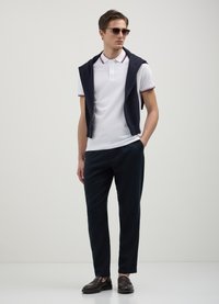 Polo bianco con colletto a due bottoni, maglione blu scuro drappeggiato sulle spalle, pantaloni blu scuro e mocassini marrone scuro.