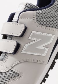 Grå lädersneaker med nätpaneler, försedda med kardborreband och en vit logo "N." Texturerade accenter förbättrar hållbarhet och stil.