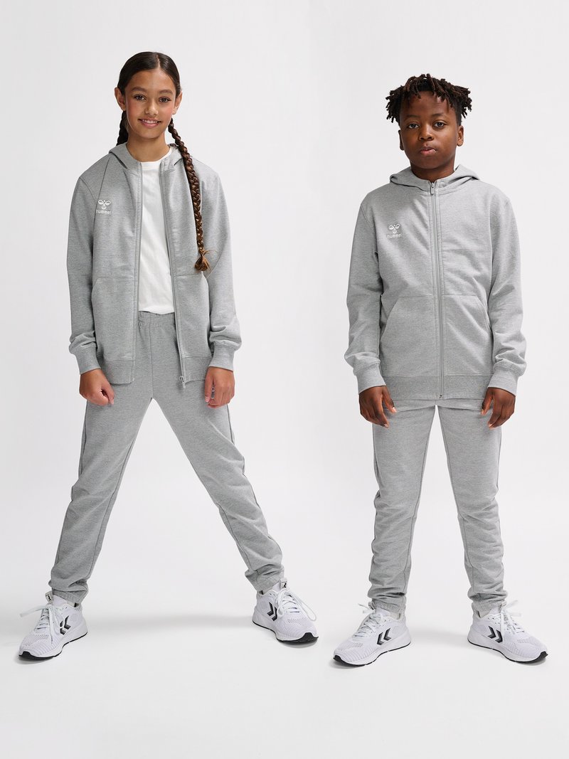Zwei Kinder, ein Mädchen und ein Junge, tragen passende graue Kapuzenanzüge und weiße Turnschuhe und stehen vor einem schlichten Hintergrund.