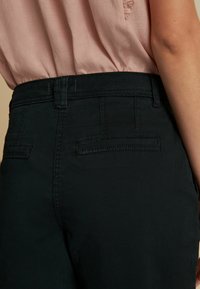 Sorte bukser med en glat stofoverflade, har en tætsiddende talje, baglommer og et klassisk design. Kontrastsyninger er synlige.