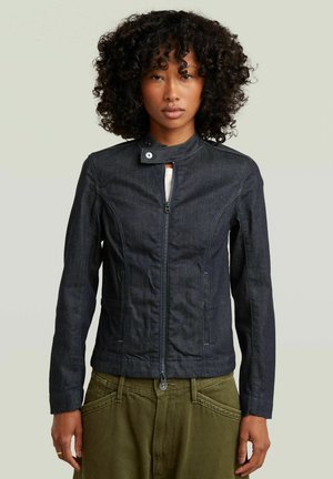 Donna con capelli ricci che indossa una giacca con zip in denim scuro e pantaloni verde oliva, in piedi davanti a uno sfondo chiaro e uniforme.