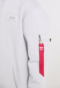 Felpa grigia con cappuccio, dotata di un'etichetta rossa rimovibile attaccata da una cerniera argentata, con un patch logo testurizzato sulla parte superiore del braccio e polsini a coste.