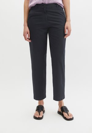 Vrouw gekleed in zwarte rechte broek, zwarte sandalen met open tenen en een paarse gebreide top, staand tegen een effen witte achtergrond.