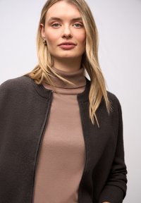 Tmavě šedý kardigan s kulatým výstřihem, texturovanou látkou a celým zipem, nošený přes přiléhavý hnědý rolák. Hladký a elegantní design.