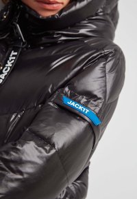 Czarna kurtka puchowa o błyszczącej teksturze, wysoki kołnierz i niebieska naszywka z napisem "JACK1T" na lewym rękawie.