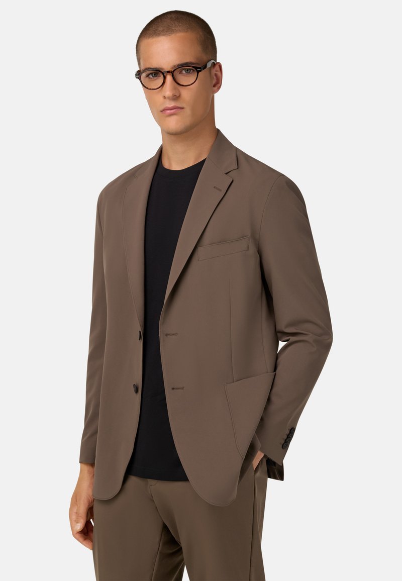 Ruskea räätälöity bleiseri, joka on valmistettu sileästä kankaasta. Siinä on leikkuriolka, kaksi nappia ja yksi tasku. Päällä mustan paidan kanssa.