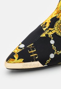 Melna auduma potīte ar dzeltenu ziedu un ķēdes rakstu, ar smailu pirkstu un zelta logo akcentu uz papēža. Elastīgs materiāls, lai nodrošinātu piemērotību.