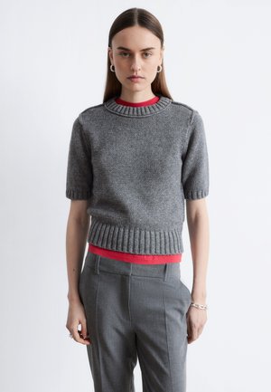 Grauer gestrickter Pullover mit kurzen Ärmeln und geripptem Ausschnitt sowie Saum, über einem roten Hemd getragen. Kombiniert mit grau geschnittenen Hosen.