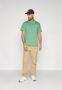 Λαχανί κοντομάνικη μπλούζα και μπεζ παντελόνι cargo, και τα δύο με το λογότυπο The North Face. Φορεμένα με μαύρο καπέλο και λευκά αθλητικά παπούτσια.