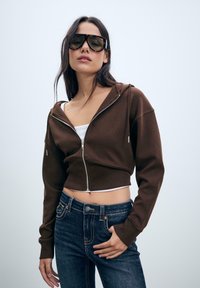 Hnedá skrátená mikina na zips vyrobená z mäkkého materiálu, má dlhé rukávy a kapucňu. Kombinovaná s tmavými džínsami a bielou tielkom.