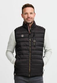 Schwarze Steppweste mit hohem Kragen, Reißverschlusstaschen und einem Abzeichen-Detail. Weiche, gesteppte Textur; über einem hellen Pullover getragen.
