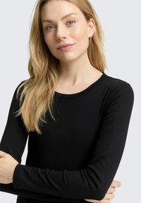 Schwarzes langärmliges Shirt aus einem weichen, dehnbaren Material mit rundem Ausschnitt und schmalen Ärmeln, getragen von einem Modell mit langen Haaren.
